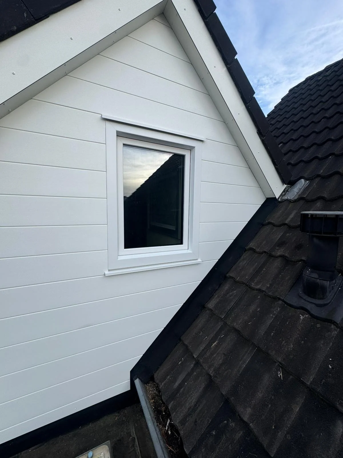 Close-up view of a modern house exterior showing a white vinyl-sided gable wall with a small window and a black tiled roof with a chimney.