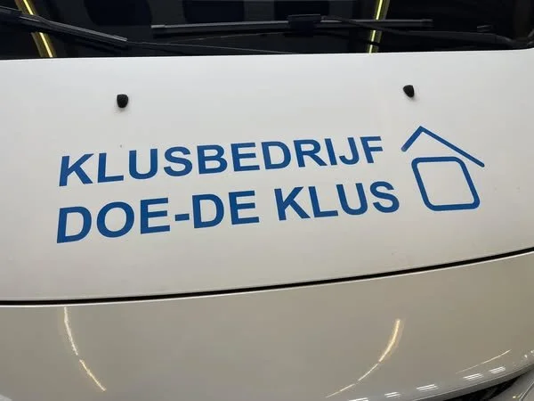 The image shows the logo and name of a club in Dutch, 'Klusbedrijf Doe-De Klus,' with a simple house icon, printed on the front of a white vehicle.
