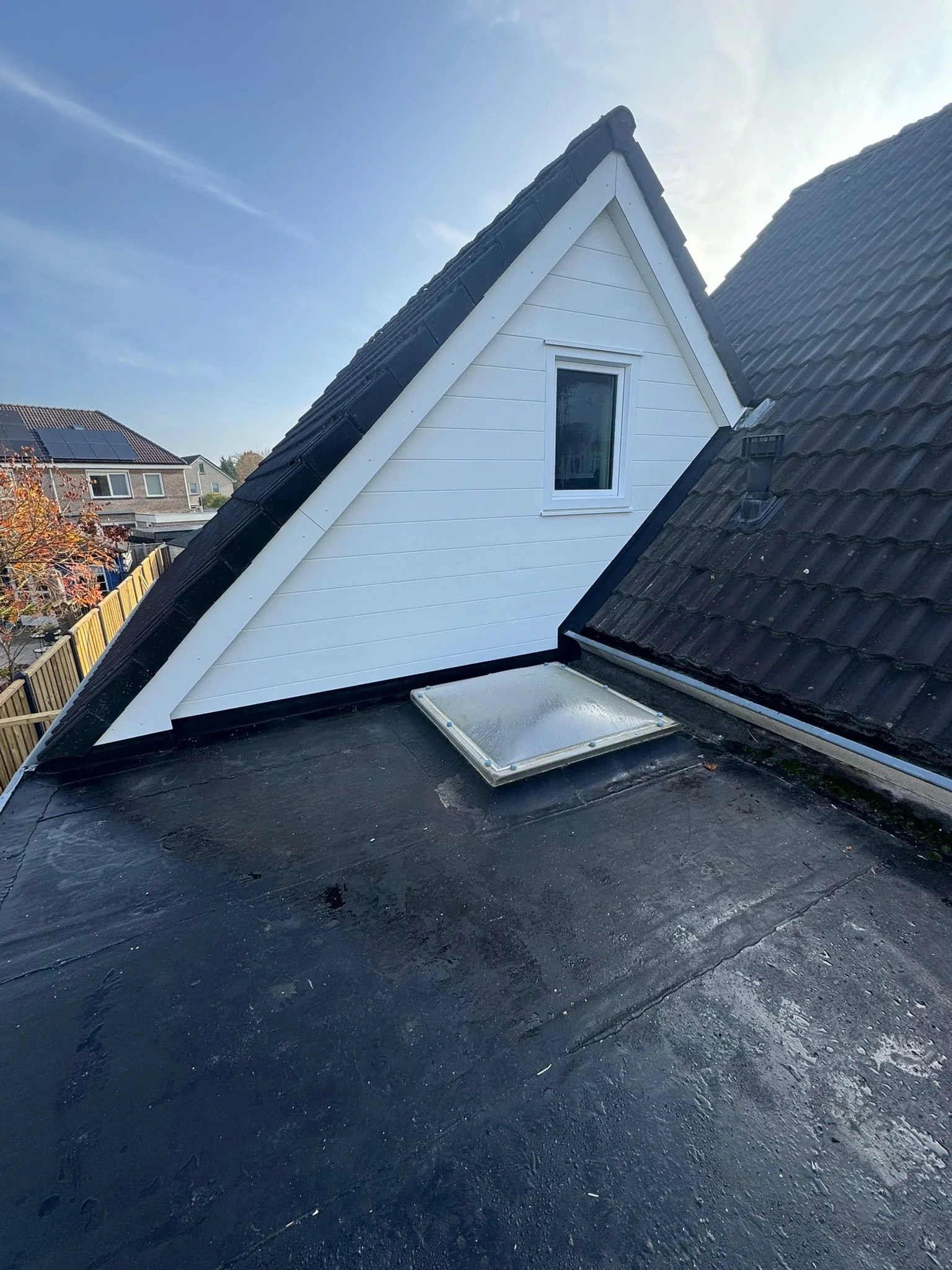 View of a rooftop with a skylight window and a white house with black roof tiles in the background, under a clear blue sky.