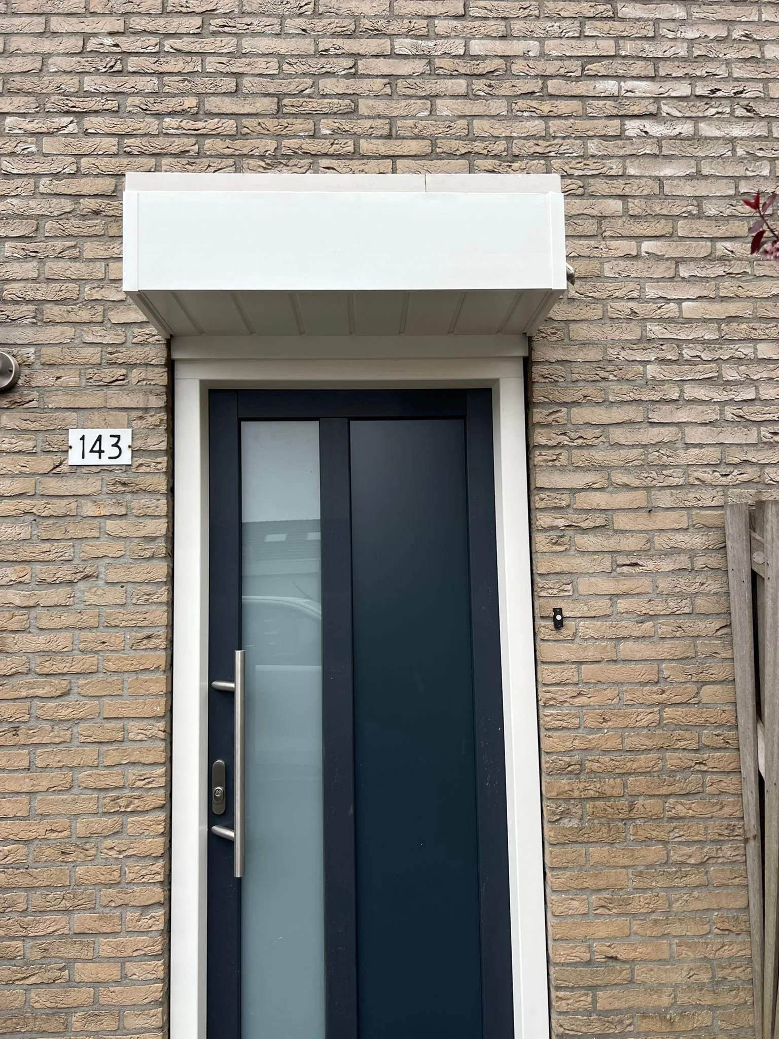 Close-up of a modern brick house entrance with a black door that has a frosted glass panel, a vertical stainless steel handle, and house number 143 on the wall.