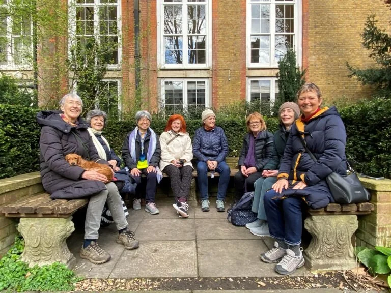 A group of 7 people sit in the benches of the Southwood Garden at St James Piccadilly, just before the Eco-contemplative liturgy service starts.  All are wearing warm coats and are smiling. A small dog rests on one woman's lap.