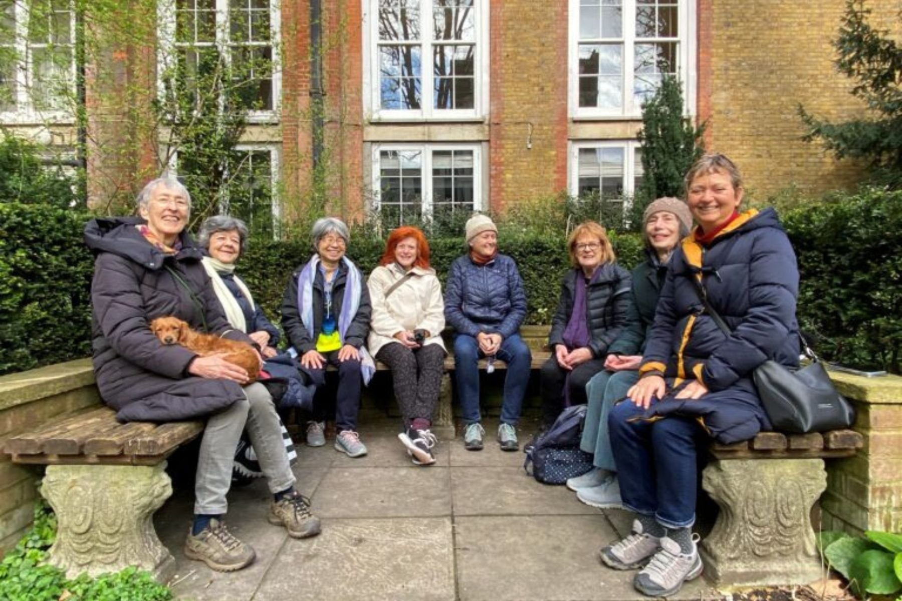 A group of 7 people sit in the benches of the Southwood Garden at St James Piccadilly, just before the Eco-contemplative liturgy service starts.  All are wearing warm coats and are smiling. A small dog rests on one woman's lap.