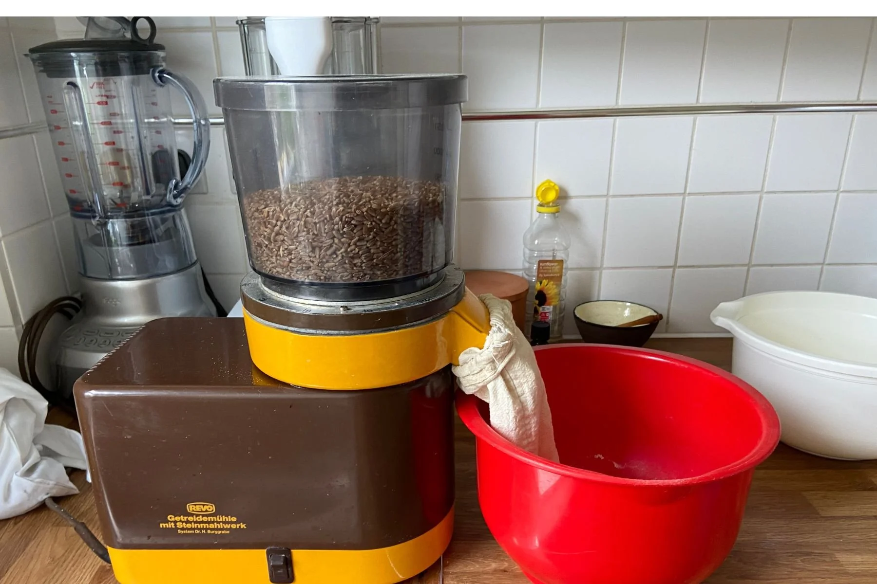 A yellow grain mill stands on a kitchen worktop. It is processing wheat grains. The processed grains are being transferred into a red mixing bowl.