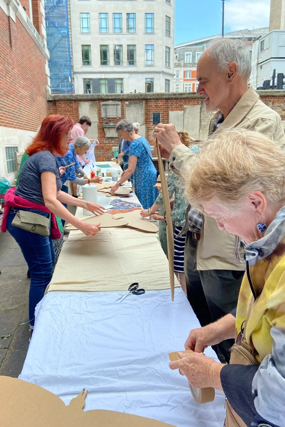 A group of people are standing at a long table in the St James Piccadilly courtyard. They are making large cardboard butterfly shapes as part of the SWARM project.
