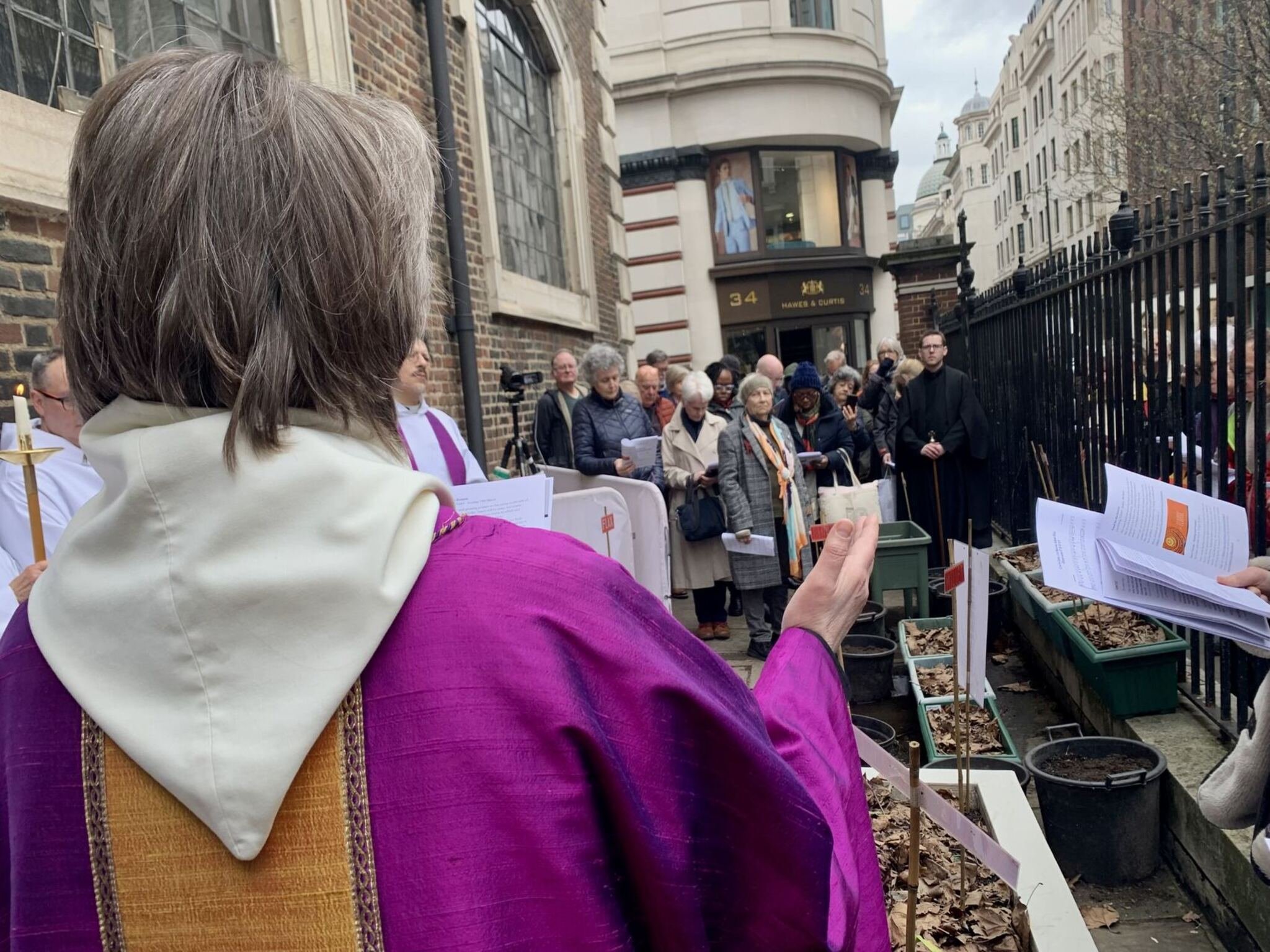 A person in a purple and cream robe leads a gathering outside a building with many people listening and holding papers.