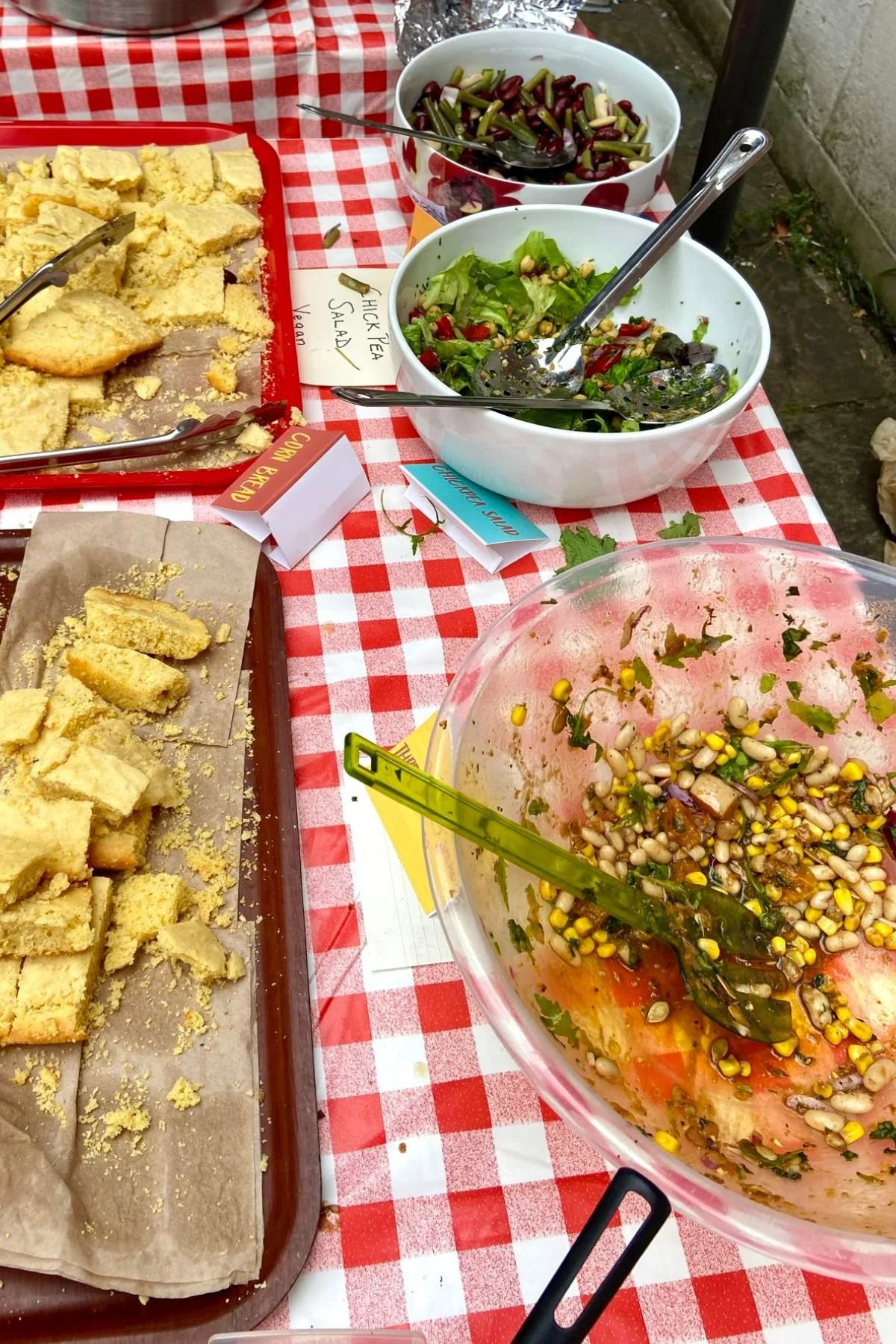 A variety of salads, beans and cornbread on a red and white checkered tablecloth, including a green beans salad, a mixed greens salad, and a black bean and corn salad.