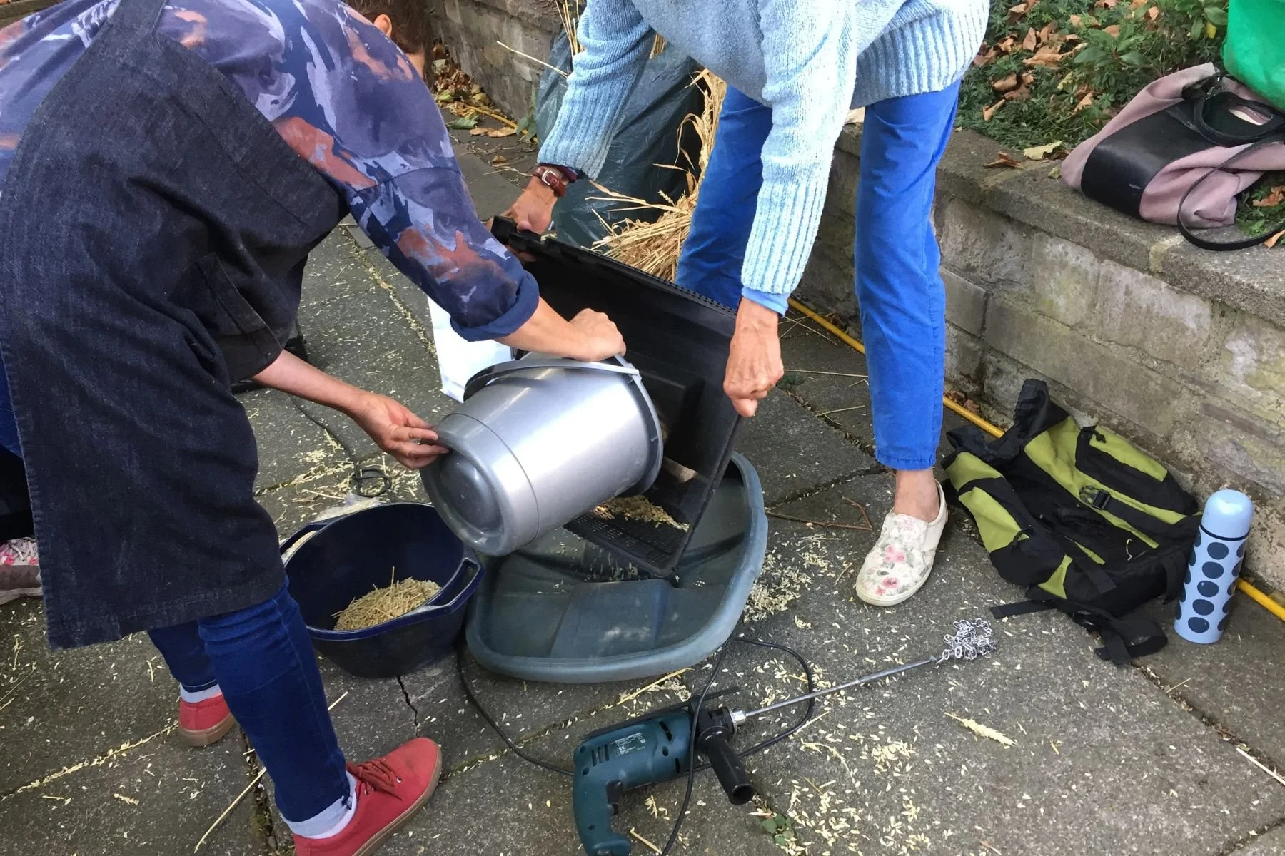 Two people stand outside and work together to process and mill wheat stalks. They transfer the stalks from one container to another.