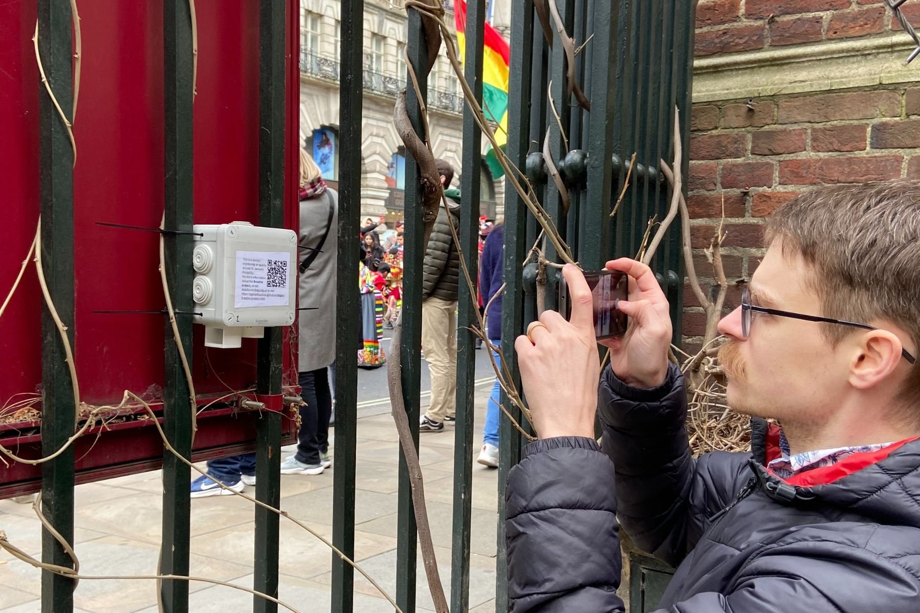 Jonathan Sutton, wearing glasses, takes a photo of a small white air quality monitoring box attached the black railings at the front of St James Piccadilly. People walking down Piccadilly are visible through the railings. 