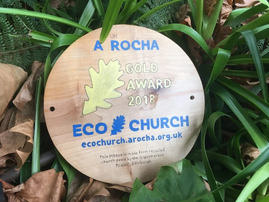 A circular wooden plaque says A Rocha Eco Church Gold Award 2018, with a gold leaf illustration. The plaque is nestled among green leaves.