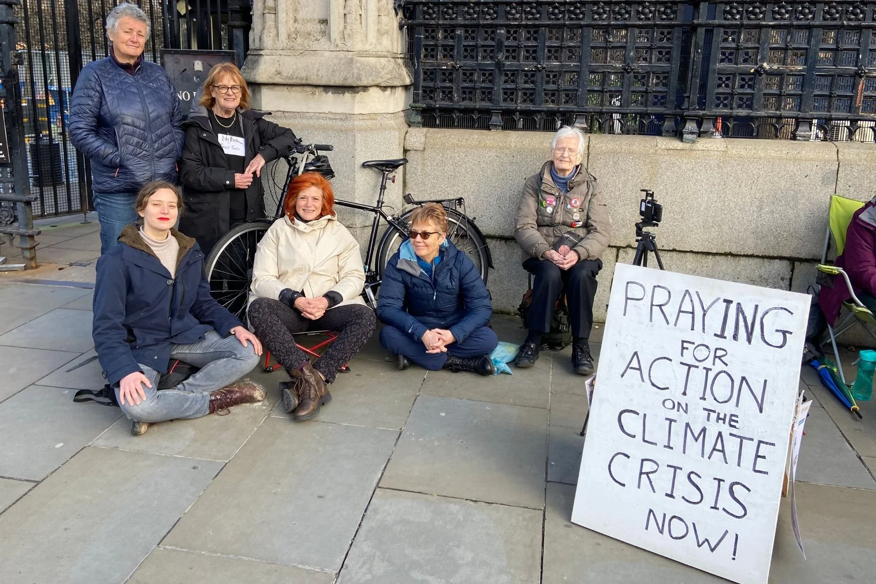 Group of seven people sit outside the Houses of Parliament in London. A handmade sign reads "Praying for action on the climate crisis now!"