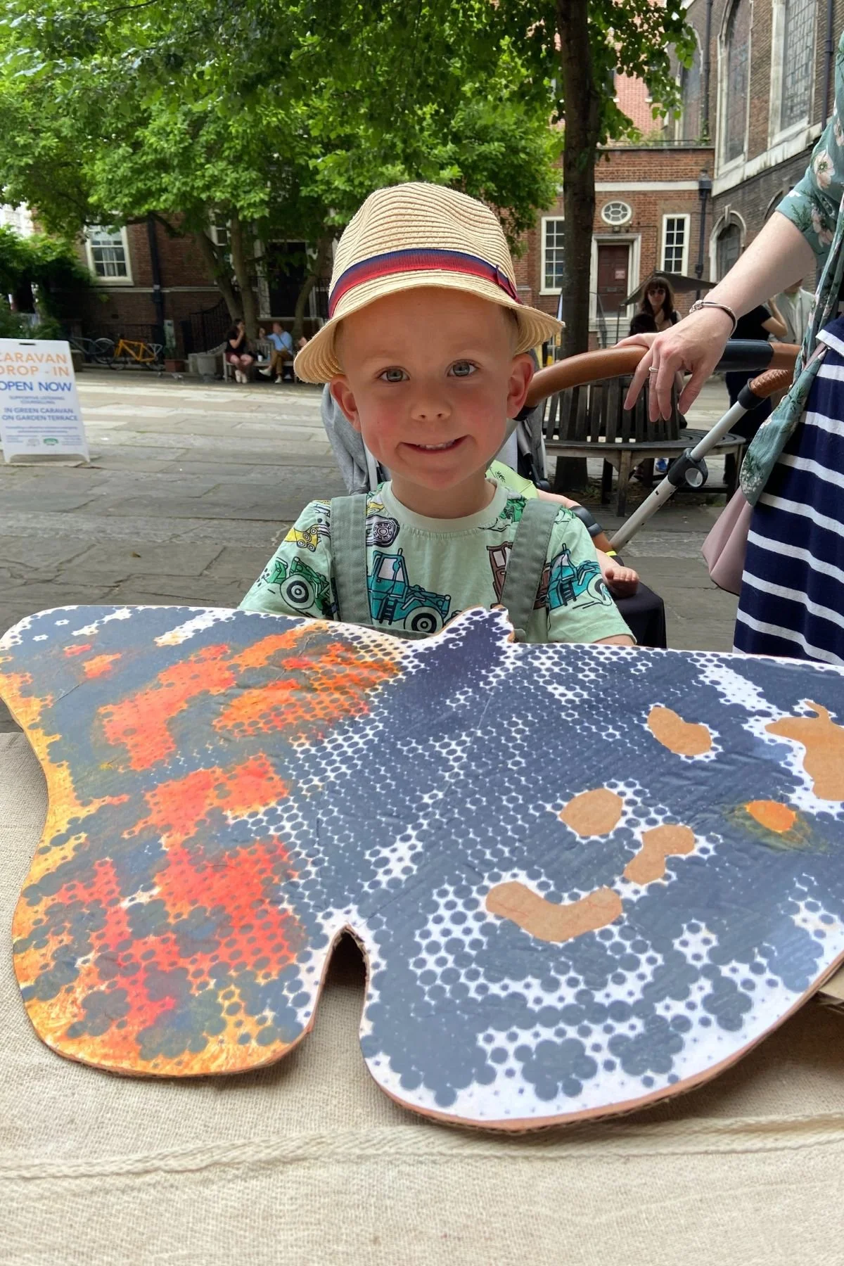 A smiling young boy wearing a straw hat and a green t-shirt. He holds up a large black and orange cardboard butterfly shape that he has painted. 