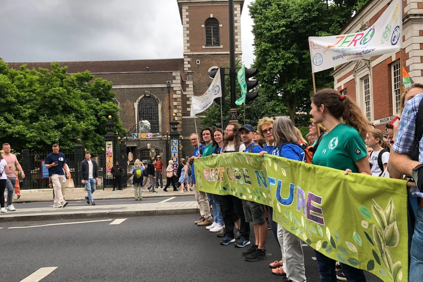 A group of people walk down Piccadilly holding a green banner that reads 'Restore Nature Now'. St James Piccadilly is visible in the background and has colourful 'Restore Nature Now' letters across the railings.
