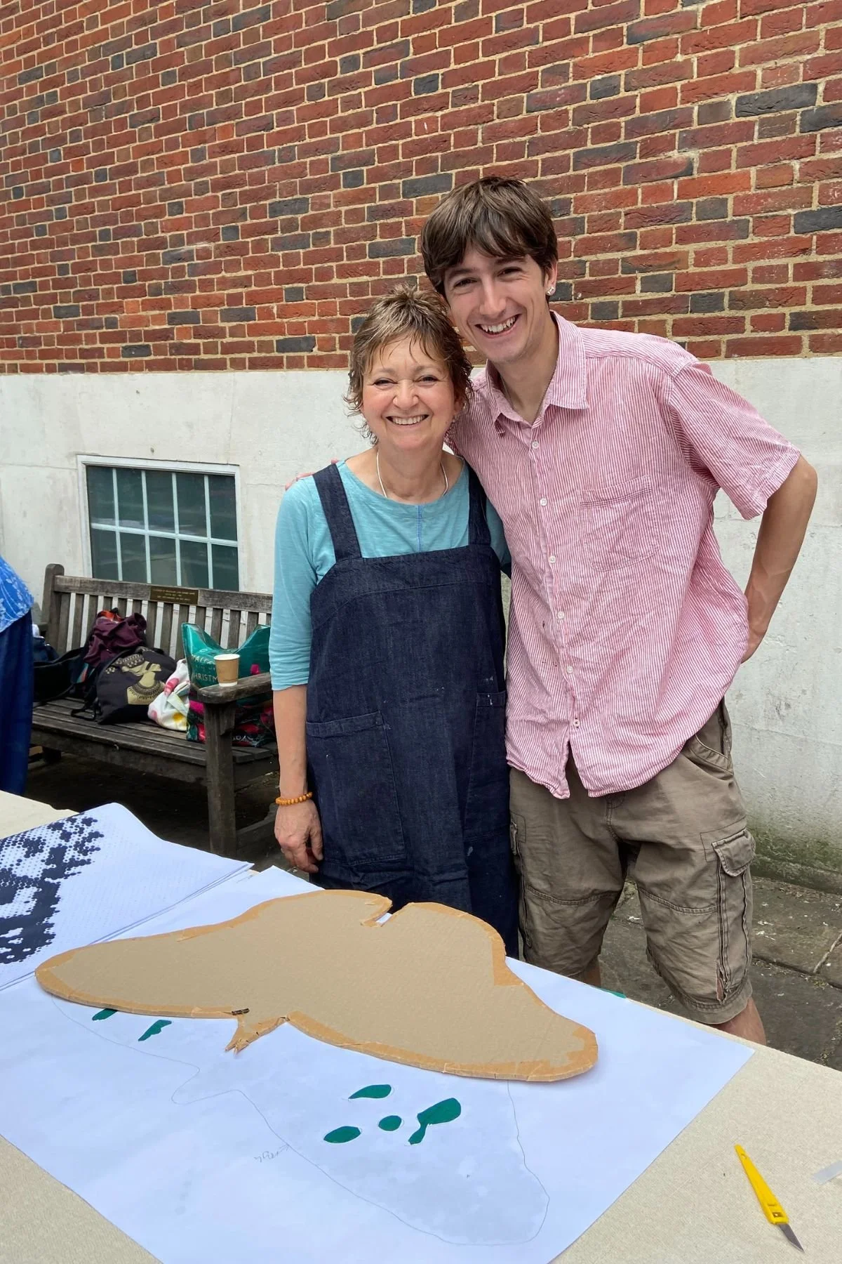 A smiling woman and a young man stand outdoors in the St James Piccadilly courtyard at a outdoors craft workshop. They are standing behind a table filled with large cardboard butterfly shapes.