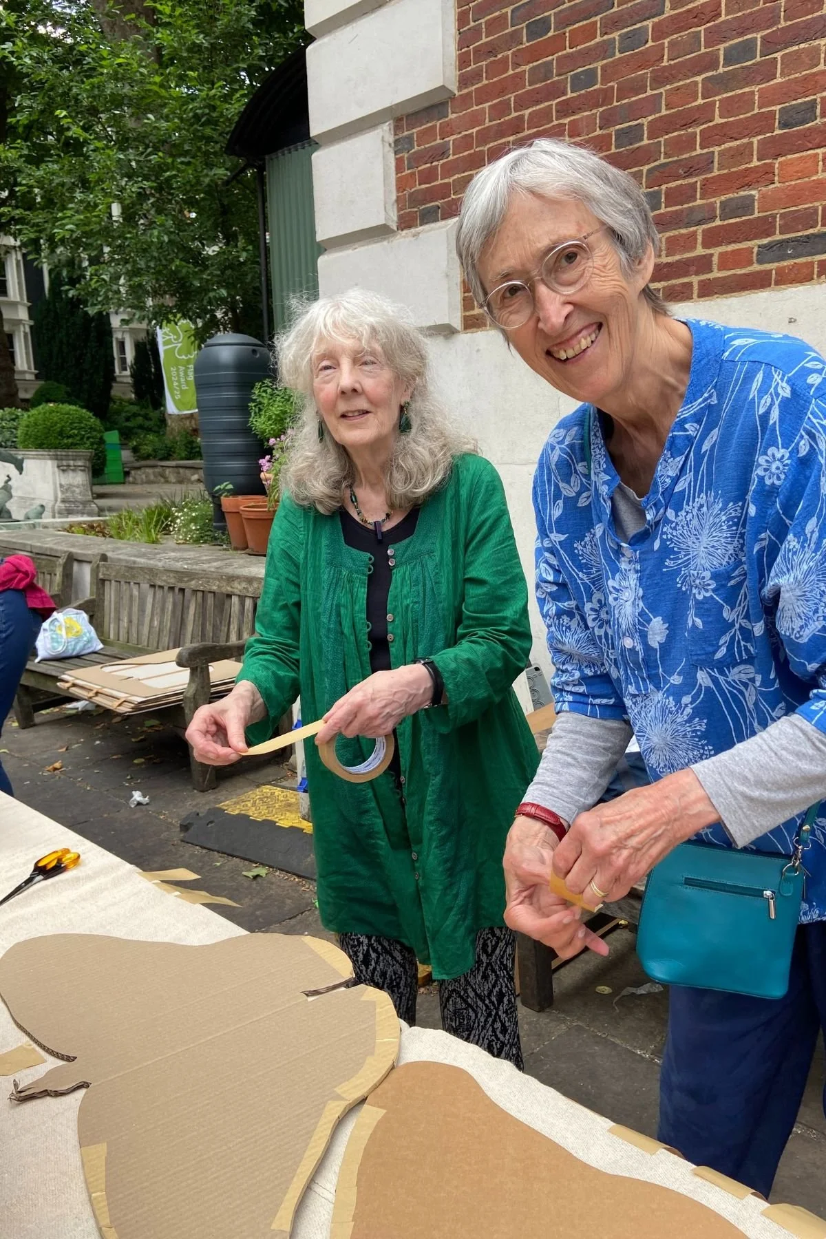Two smiling woman are standing outdoors in the St James Piccadilly courtyard. In front of them is a trestle table with large cardboard butterfly shapes. This is part of the SWARM project.