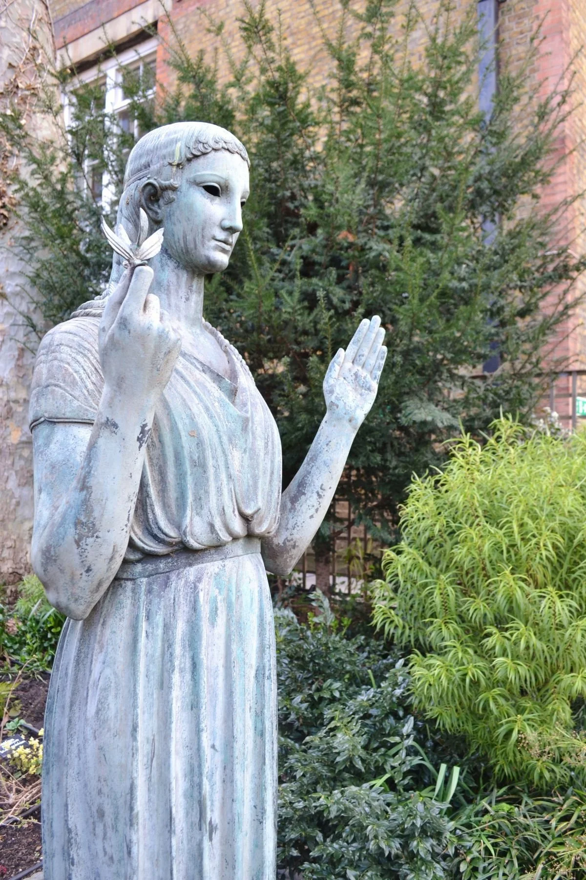 A weathered statue of a woman with a peaceful expression holding an olive branch in one hand. Her other hand is raised and open in welcome. She stands in the Southwood Garden of St James's Piccadilly. There are trees in the background.