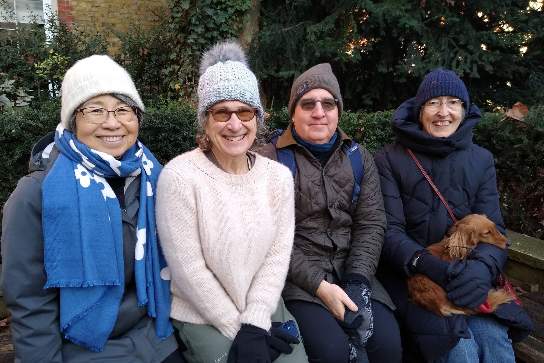 Four smiling people sitting on a bench outdoors in the Southwood garden at St James church Piccadilly. Everyone is wearing warm clothing and hats. A woman on the right is holding a small brown dog wearing a blue jacket.