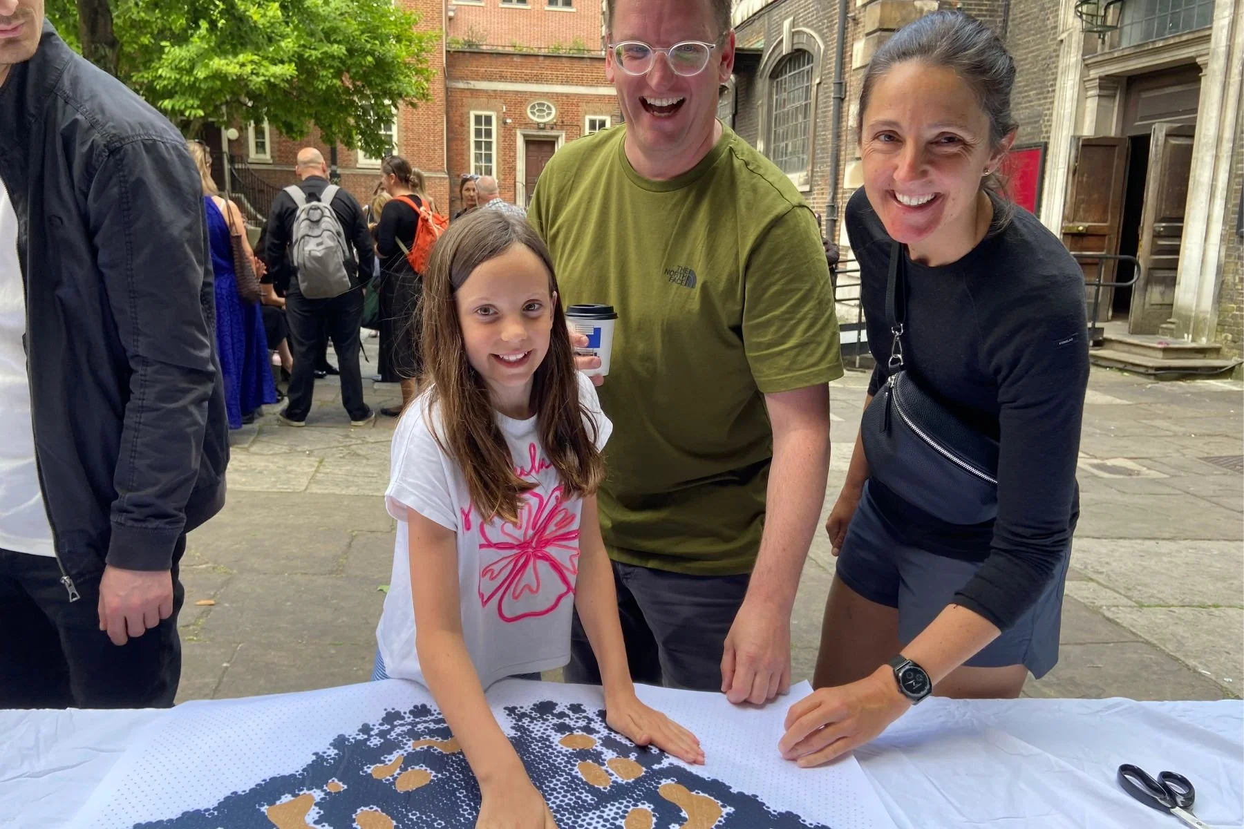 A girl and two smiling adults, a man and a woman, engaging in an arts and crafts activity outdoors with a piece of artwork on a table. The girl has long brown hair and is wearing a white shirt with a pink flower design. The man is wearing glasses and
