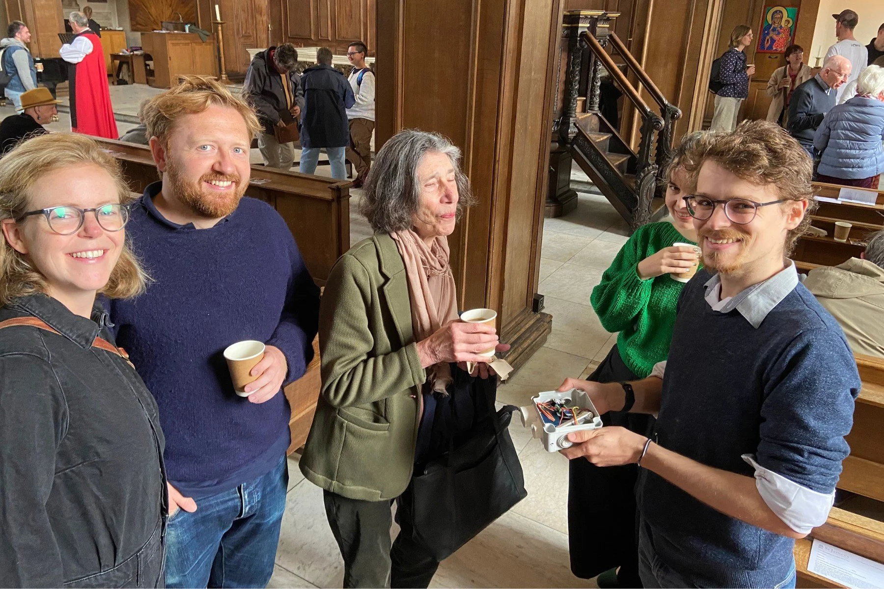 Group of five people, three women and two men, standing inside a church, smiling and holding coffee cups, with some people and a staircase in the background.