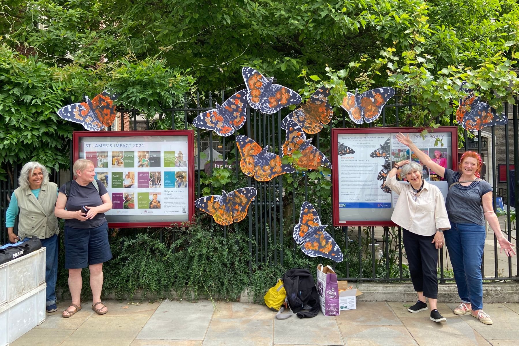 Four women stand smiling next to the SWARM butterflies art display which is attached the black railings of St James Piccadilly. There is green foliage in the background. 