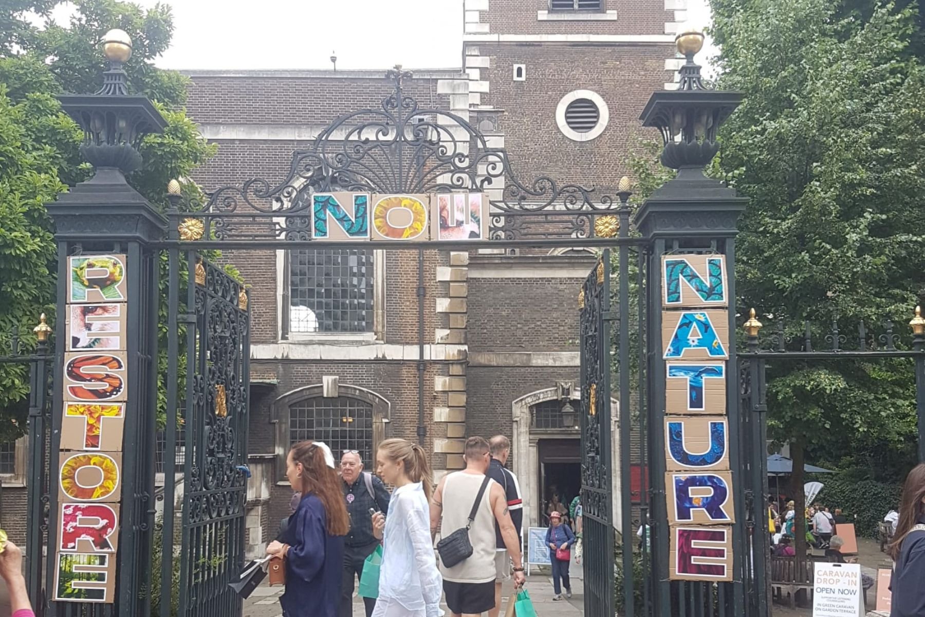 The black railings of St James' Piccadilly have colourful letters that spell 'Restore Nature now' around the open gates. People are walking through the gate and along the street.