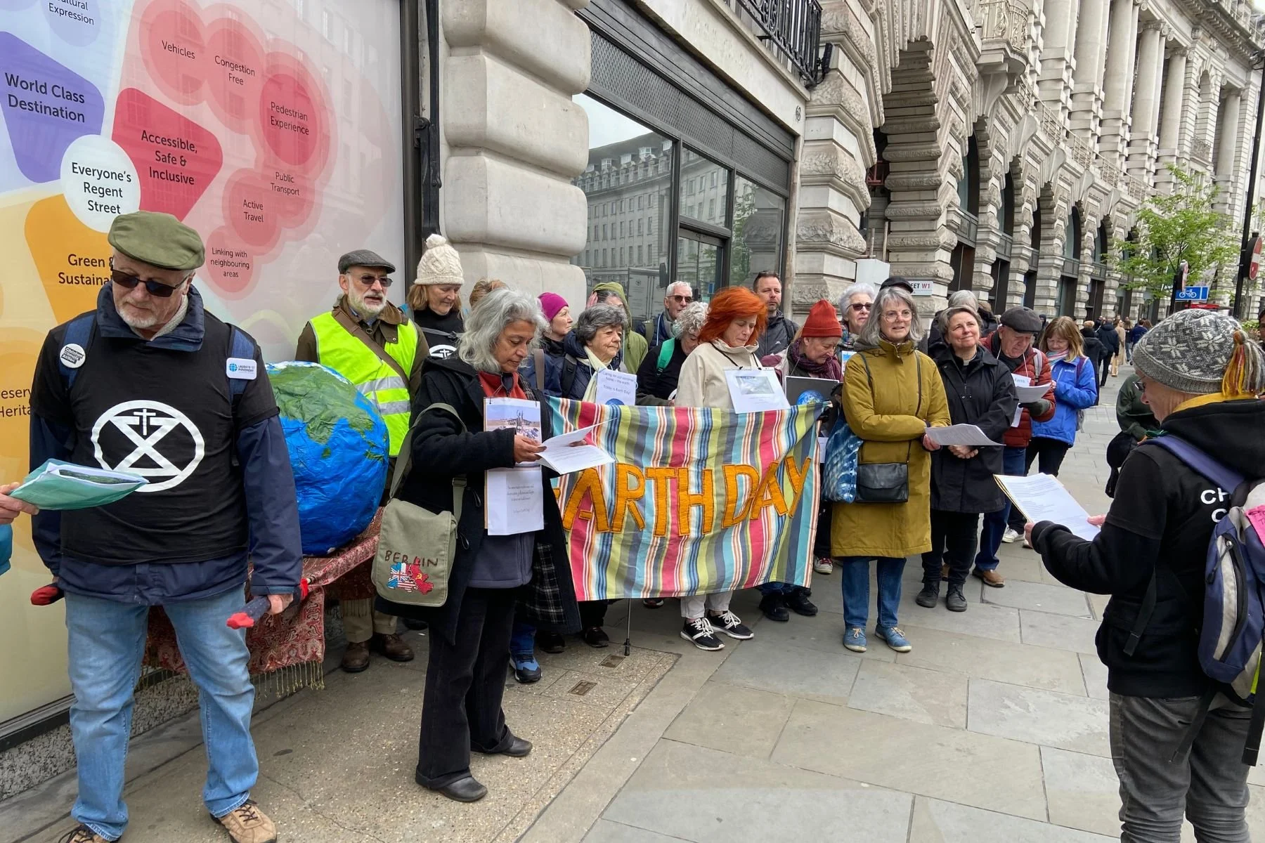 A group of people gather peacefully outside buildings on Regent Street as part of a prayer walk. They hold a colourful banner that reads 'Earth Day'. Most people are looking at the person who is speaking and leading the group.