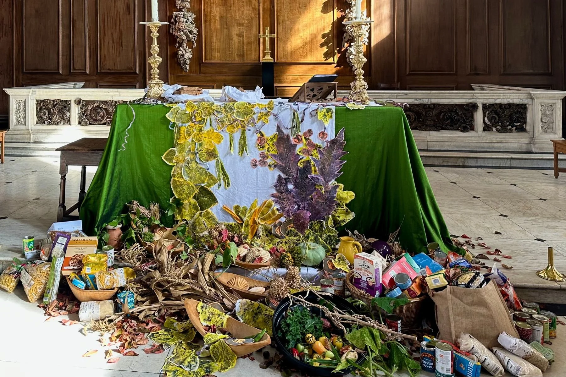 Altar decorated for the Harvest Festival at St James Piccadilly. There is a green hand printed green altar cloth. At the foot of the altar, there are fresh fruits and vegetables, autumn leaves and packaged food.
