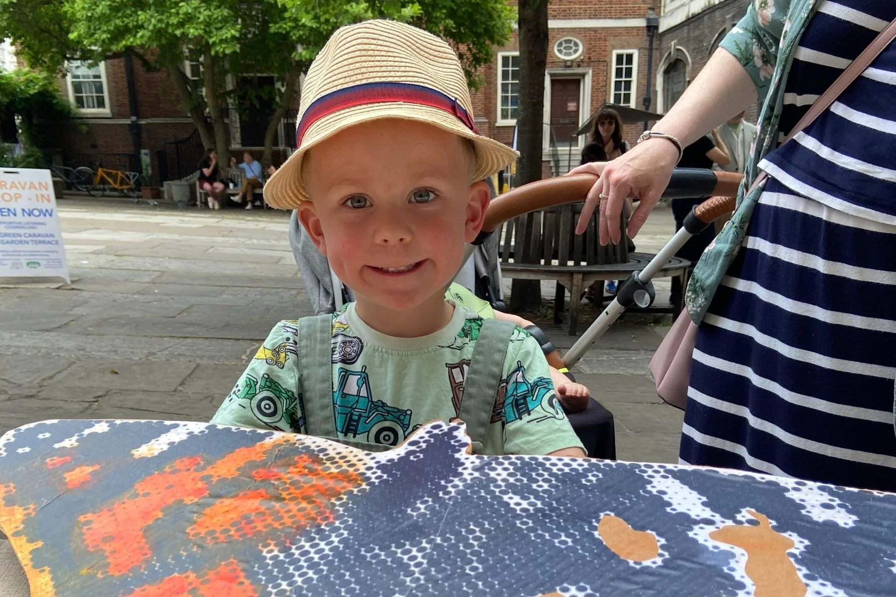 A young boy smiling at the camera, wearing a straw hat. In front of him on a table is a large black and orange cardboard butterfly shape. A woman stands next to him, just out of shot.
