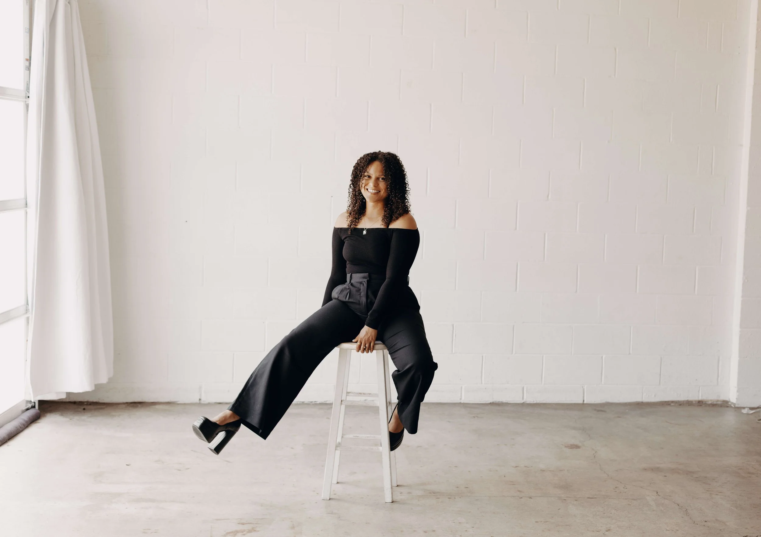 A woman with curly hair, dressed in a black off-the-shoulder top and wide-leg pants, sitting on a white stool in a minimalist room with a white brick wall background, white curtains, and natural light.