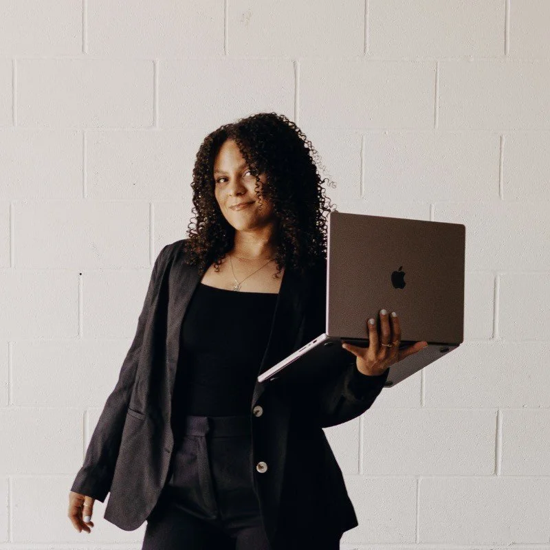 A woman with curly hair wearing a black blazer and black top holding a laptop in front of a white brick wall.