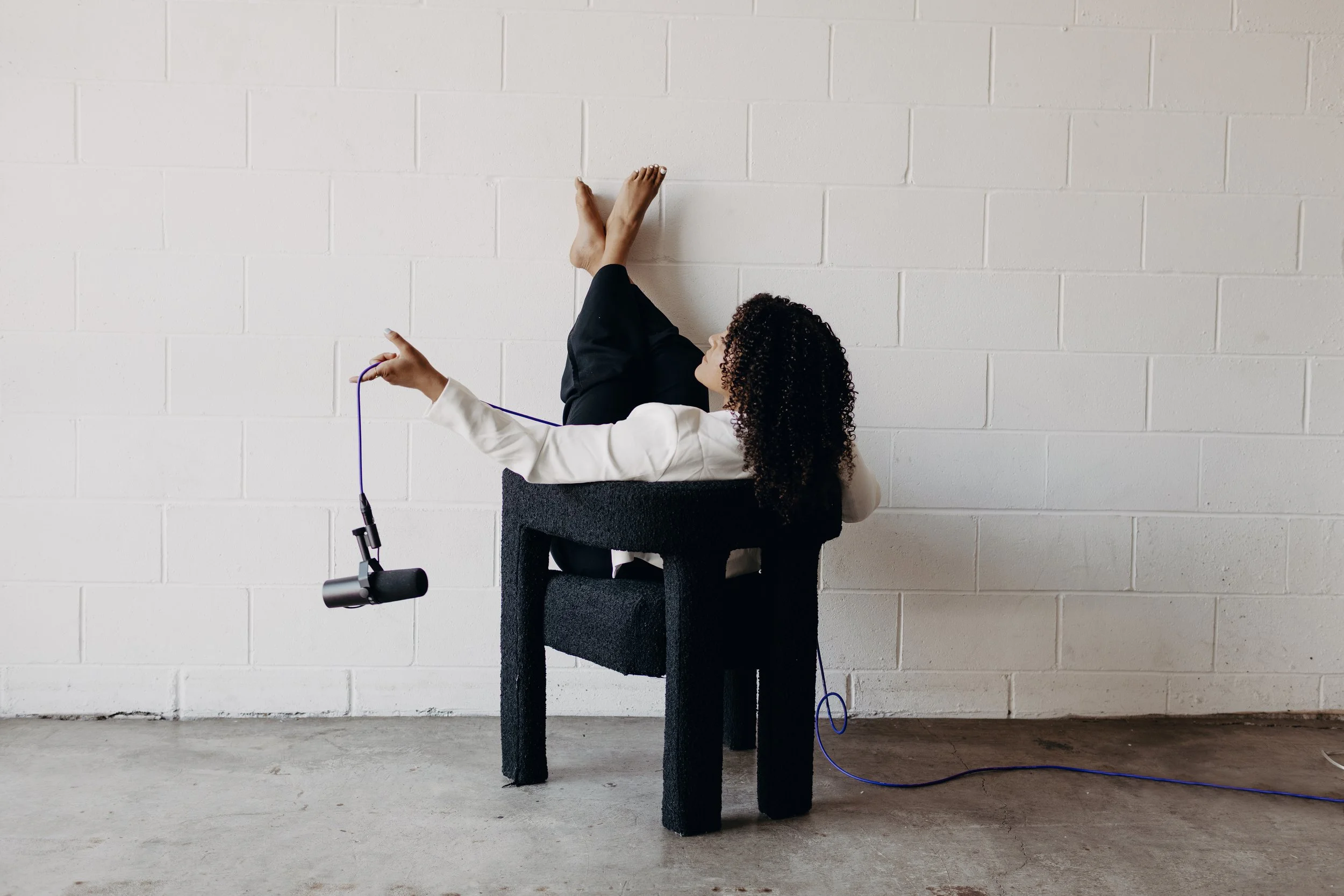 Woman with curly hair sitting on a chair with her legs up against a white brick wall, holding a purple cable connected to a microphone stand, in a minimalist room.