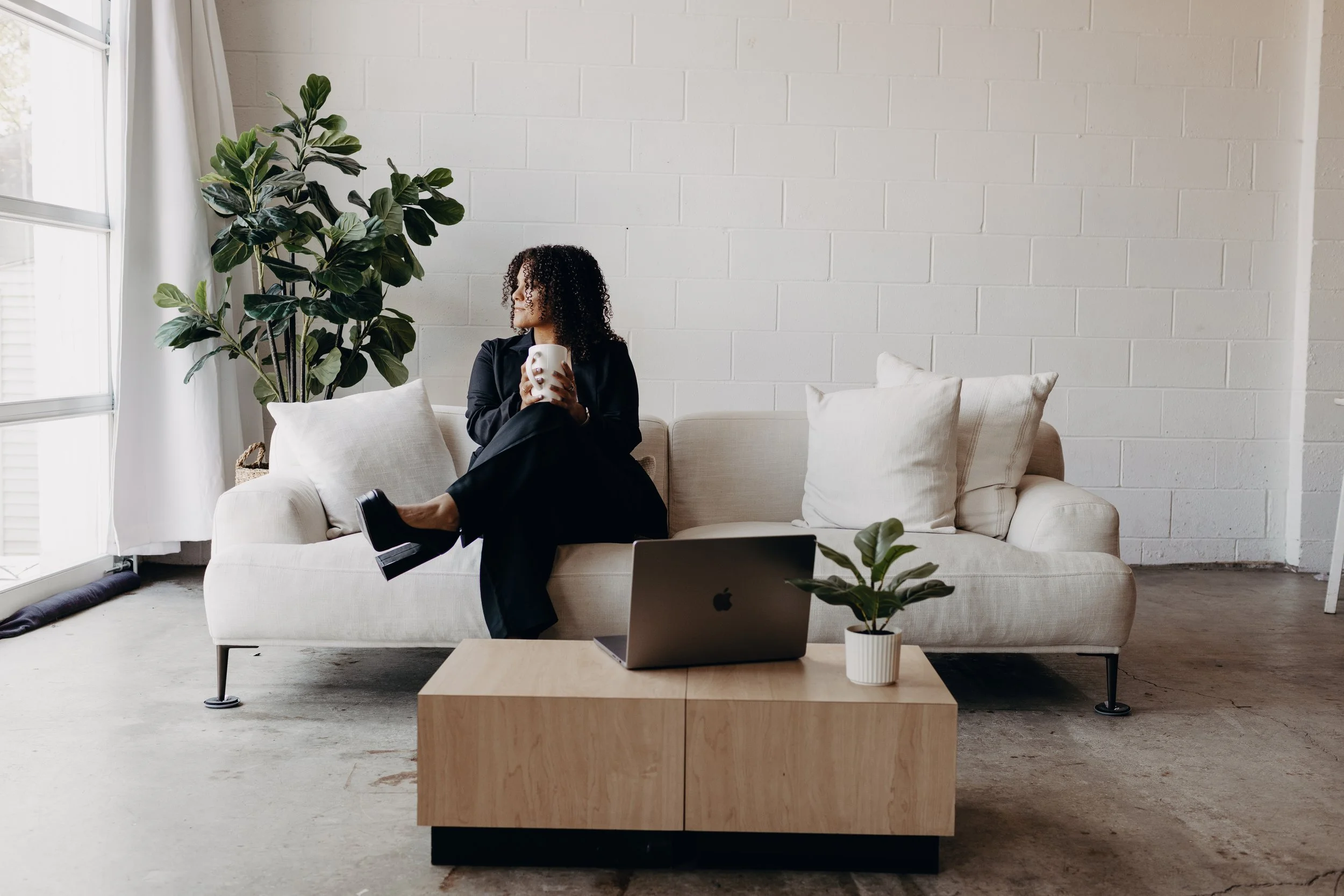 A woman sitting on a beige sofa, holding a white mug, looking out a window in a minimalist living room with plants, a wooden coffee table with a laptop and small potted plant, white pillows, and a large window with curtains.