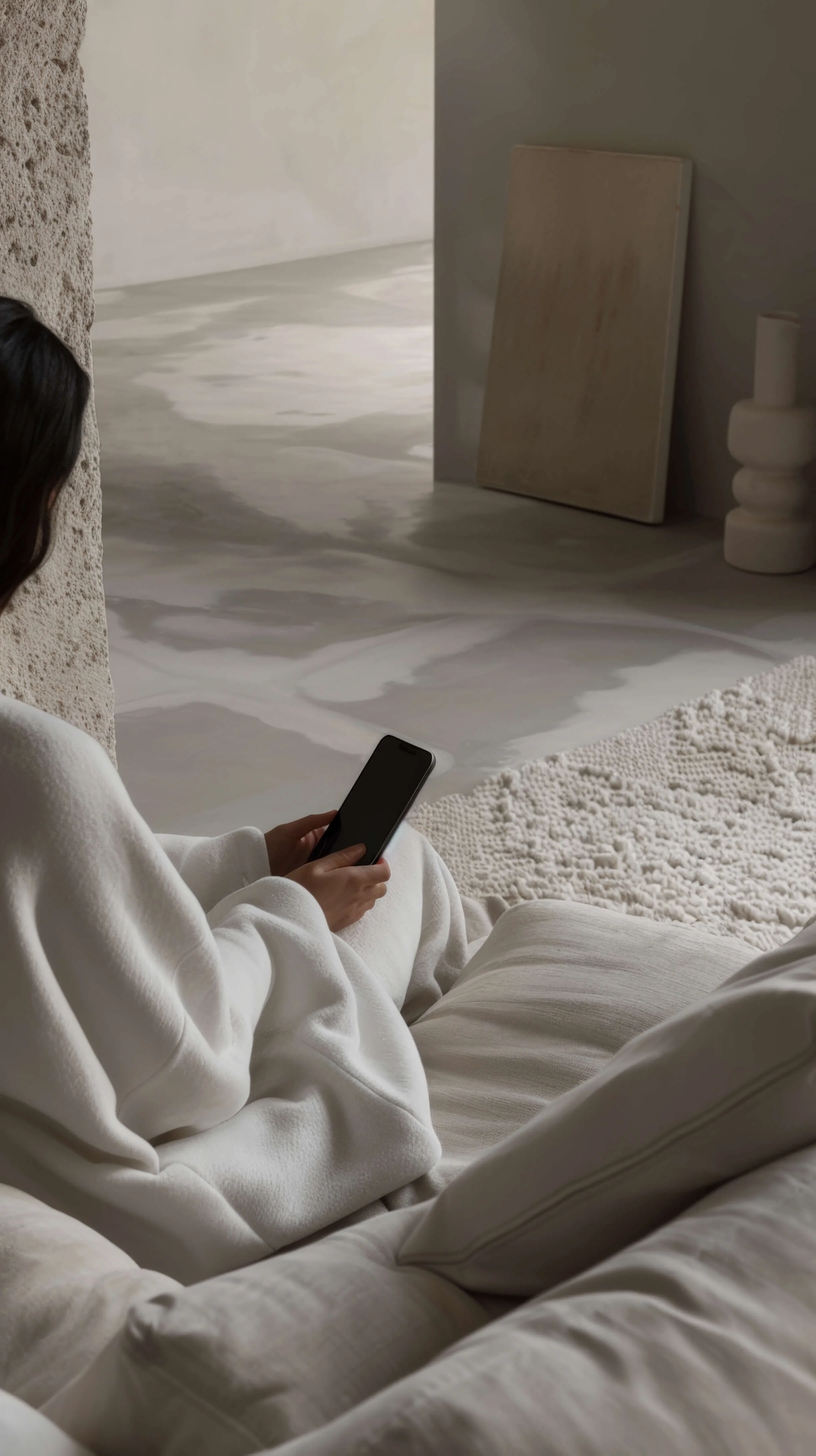 A woman in a white hoodie and beige pants sitting on a beige sofa, holding a smartphone, with a textured white rug and minimalist decor in the background.
