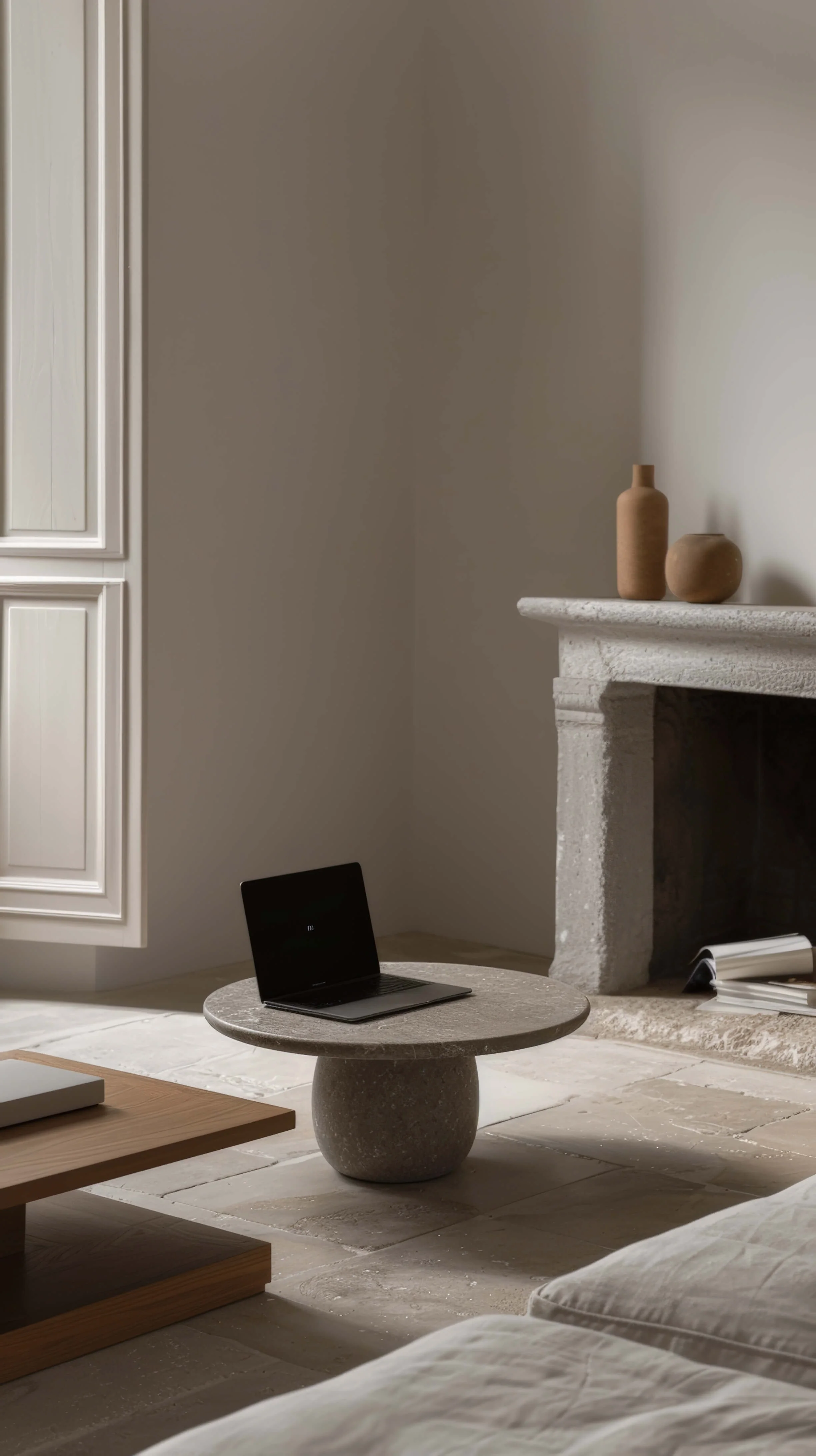 A modern living room with a stone fireplace mantel decorated with two beige vases, a small round stone table holding a black laptop, and a partially visible wooden coffee table.