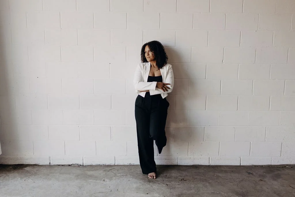 A woman with curly hair stands against a white brick wall, wearing black pants and a white blazer, crossing her arms and looking to the side.