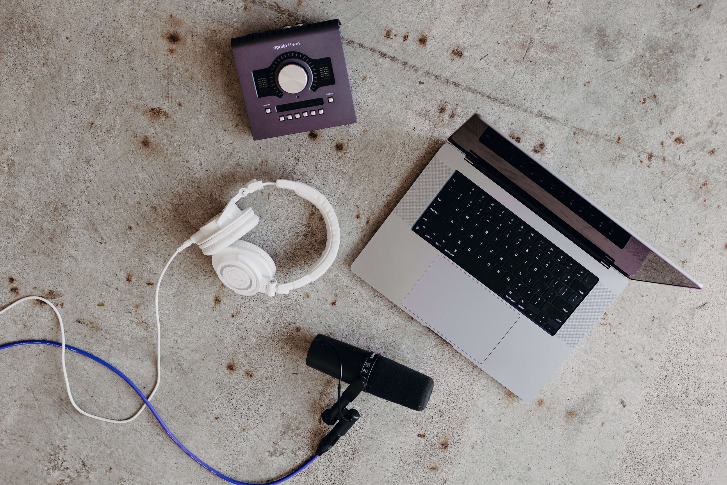 A laptop, white headphones, a microphone, and a music control device are arranged on a concrete surface.