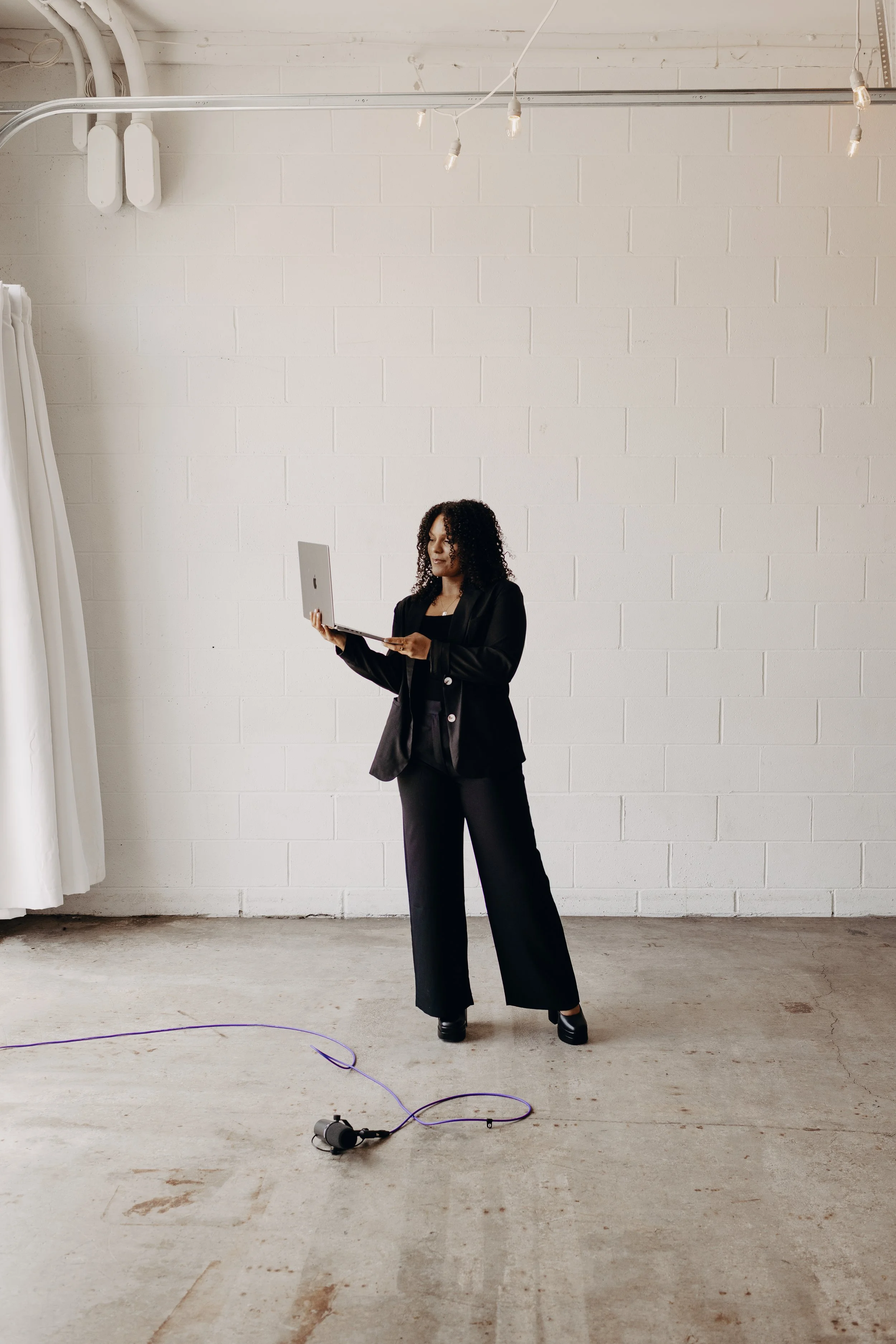 A woman dressed in black standing on a plain concrete floor, holding a laptop, in an industrial-style room with white brick walls and string lights overhead.