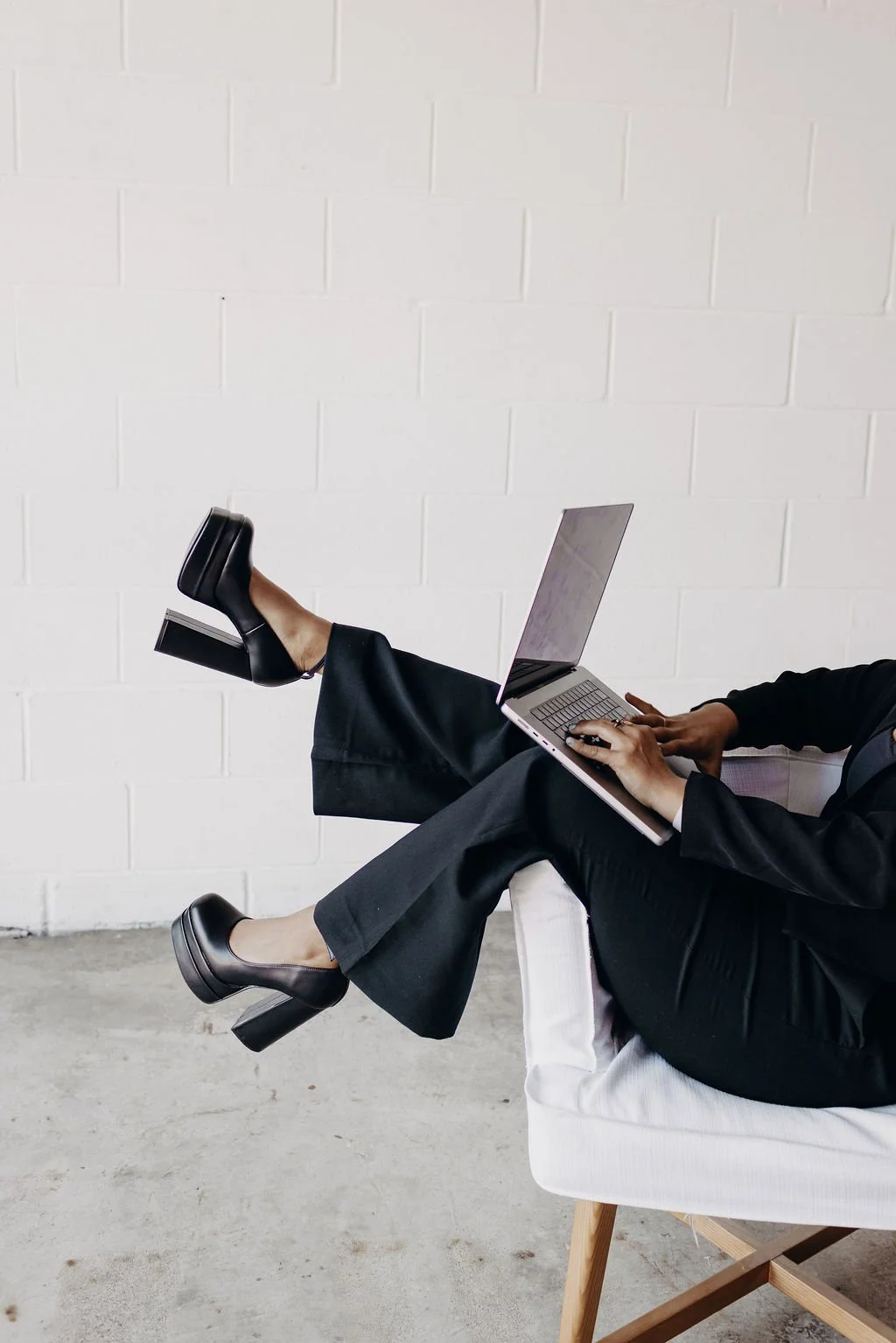 A person sitting in a chair working on a laptop with their legs crossed, wearing black high heels and black pants, against a white brick wall background.