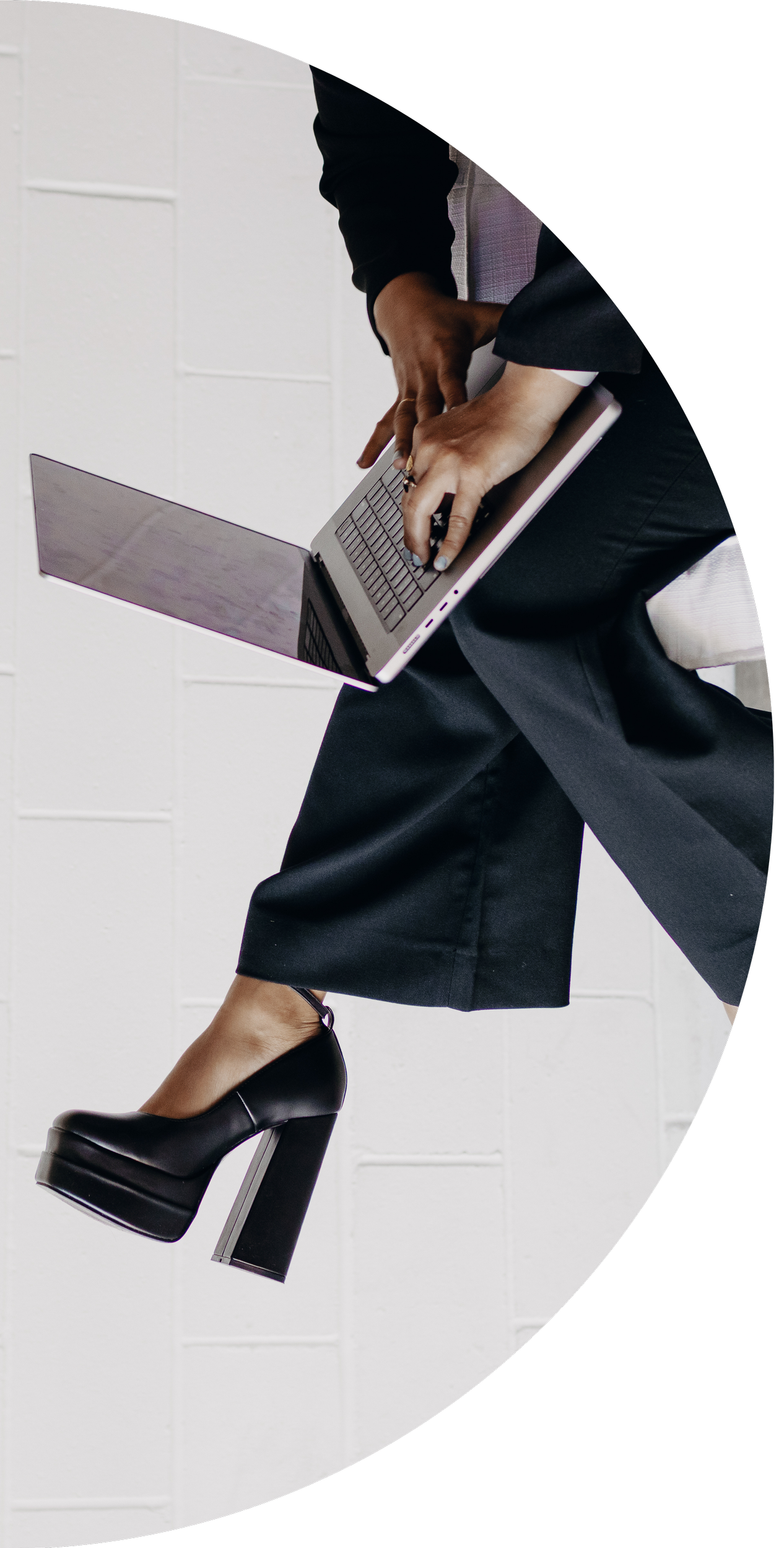 Person sitting on a chair, wearing black high-heeled shoes and dark pants, using a laptop against a white brick wall.