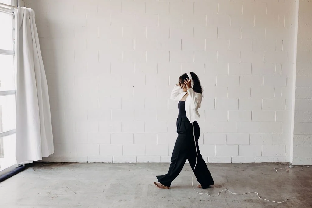 A woman with curly hair wearing a white jacket and black pants, walking barefoot in an empty room with white brick walls and a large window with white curtains, holding her head with both hands.