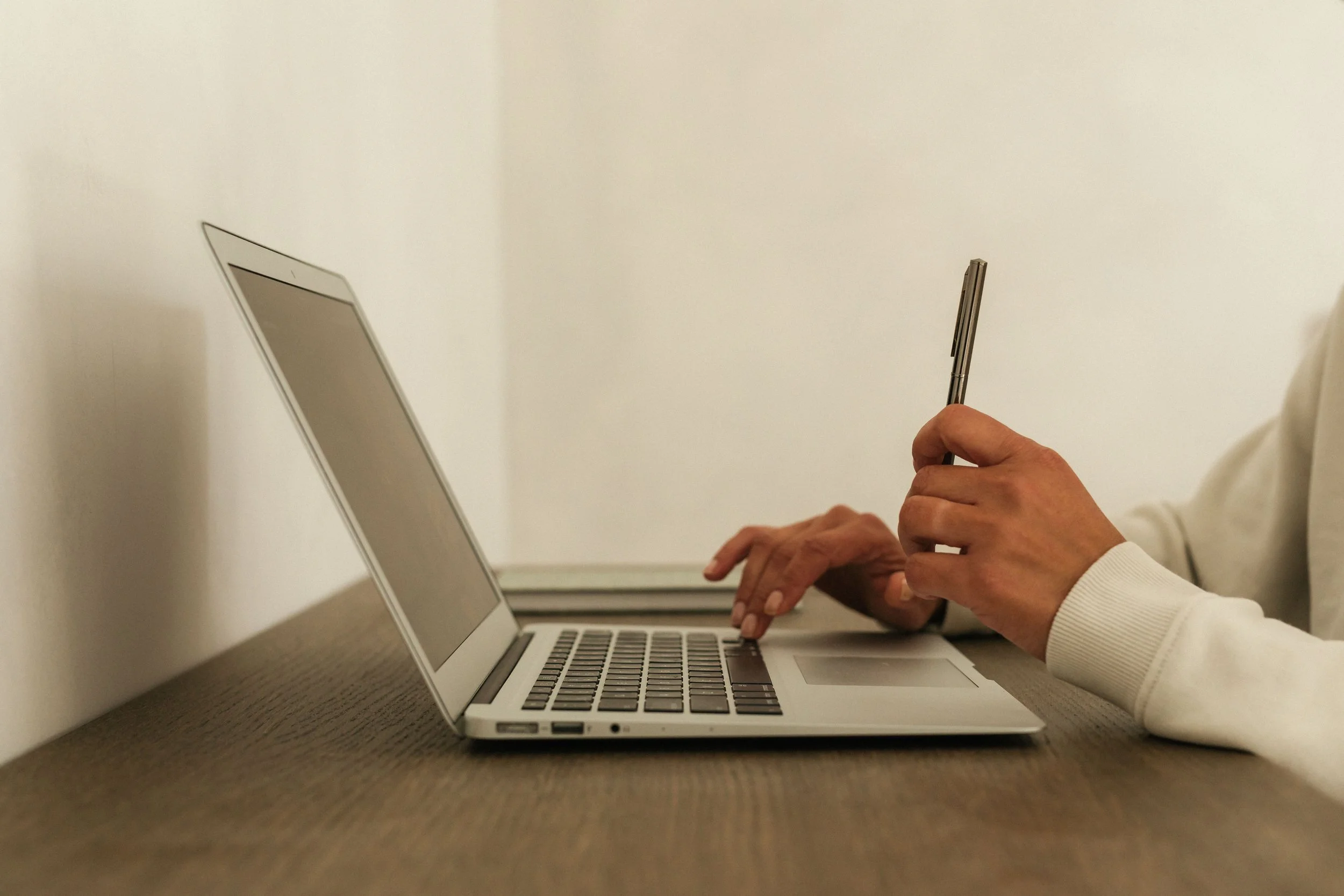 A person wearing a white shirt working on a silver laptop at a wooden desk, holding a pen in their right hand while their left hand rests on the laptop keyboard, against a plain white wall background.