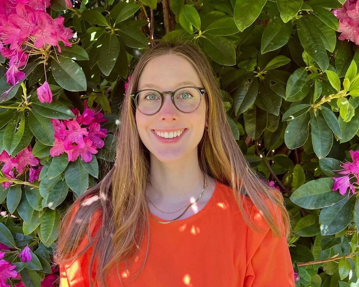 A young woman with long brown hair and glasses smiling outdoors in front of green foliage and pink flowers, wearing an orange shirt.