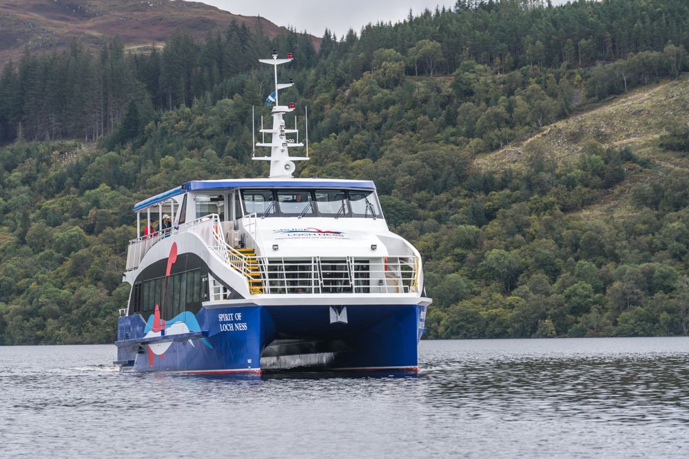 A white and blue passenger ferry boat named 'Spirit of Loch Ness' is sailing on Loch Ness with forested hills in the background.