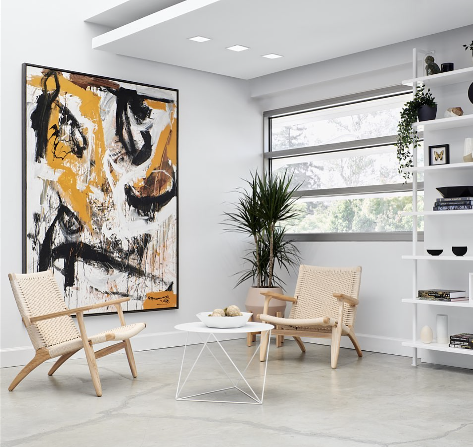 Modern living room with abstract artwork, two wooden chairs with woven seats, a white round coffee table with decorative balls, a potted plant, and white shelving with decor, books, and plants, near large horizontal windows.