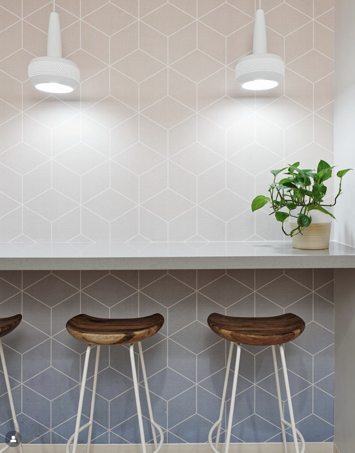 Modern office café interior with a geometric patterned tiled wall, a countertop with a potted plant, and three wooden and white metal barstools.