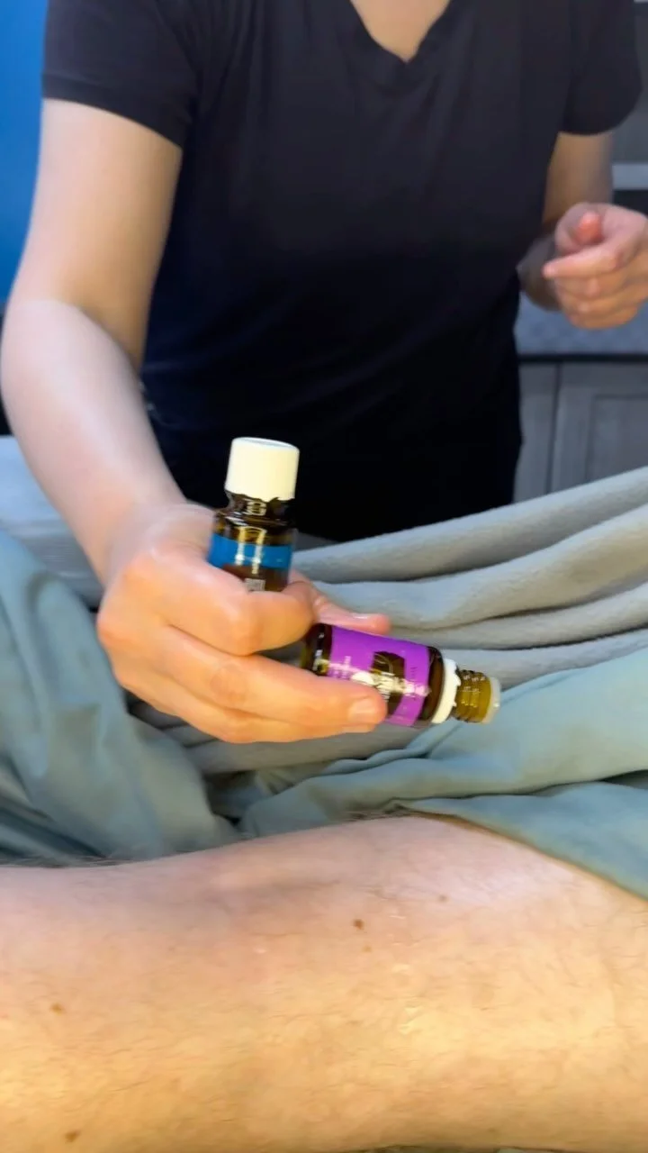 A person holding two small bottles of essential oils, preparing to apply them to a patient's leg in a clinical setting.