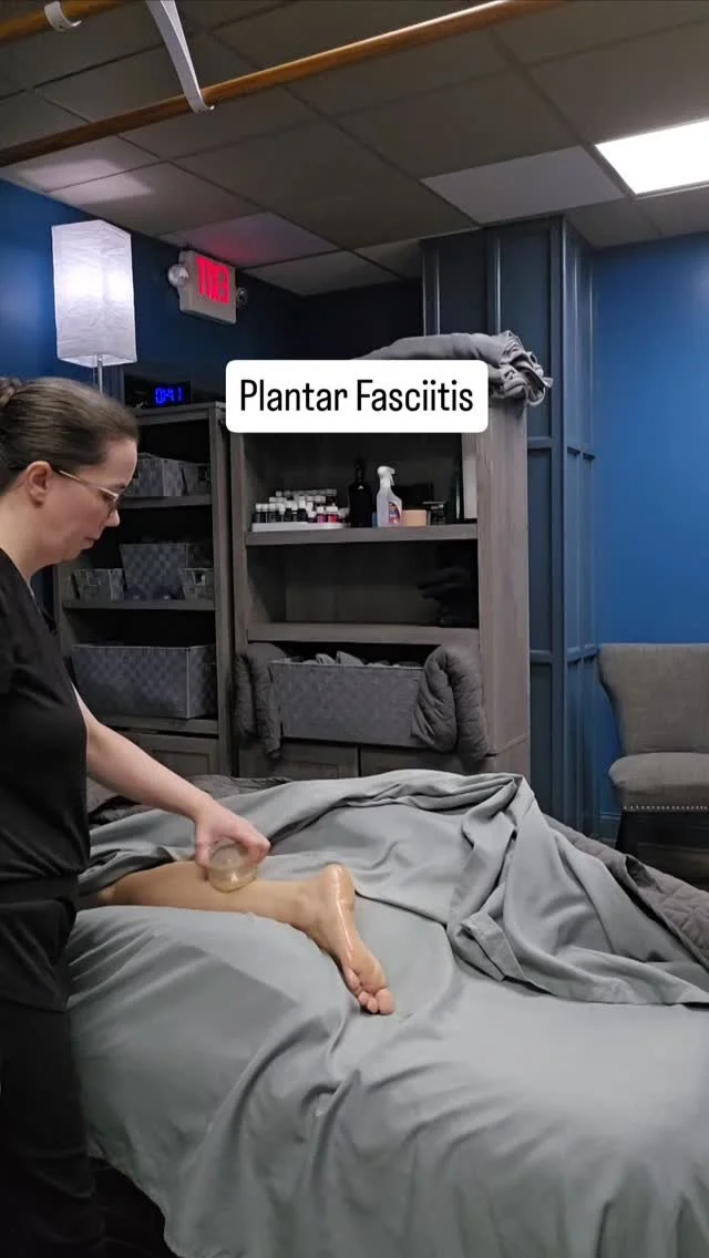 A woman in black scrubs is massaging a person's foot on a bed that is covered with gray sheets. The person lying down has their leg extended. A sign reading 'Plantar Fasciitis' is on a shelf behind them, along with various bottles and rolled-up towels in a medical or therapy room.