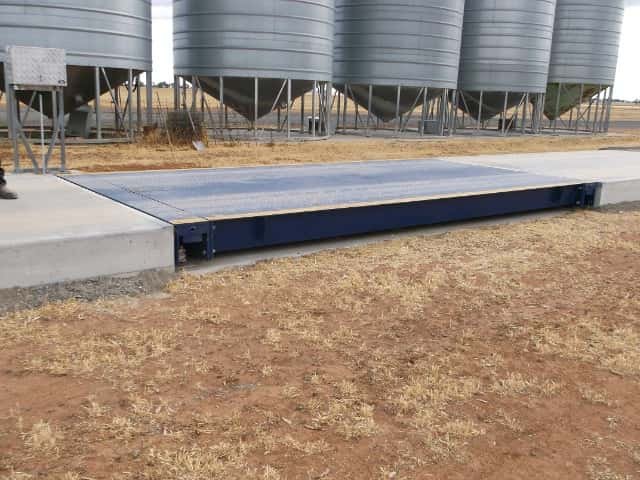 A concrete scale embedded in the ground outside, with metal grain silos in the background on a farm.