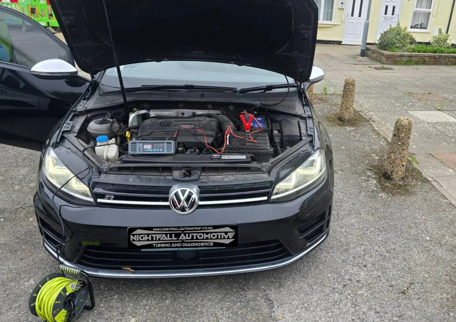A black Volkswagen car with open hood showing engine and diagnostic equipment connected, parked on a street in front of pavement and small posts, with a yellow building in the background.