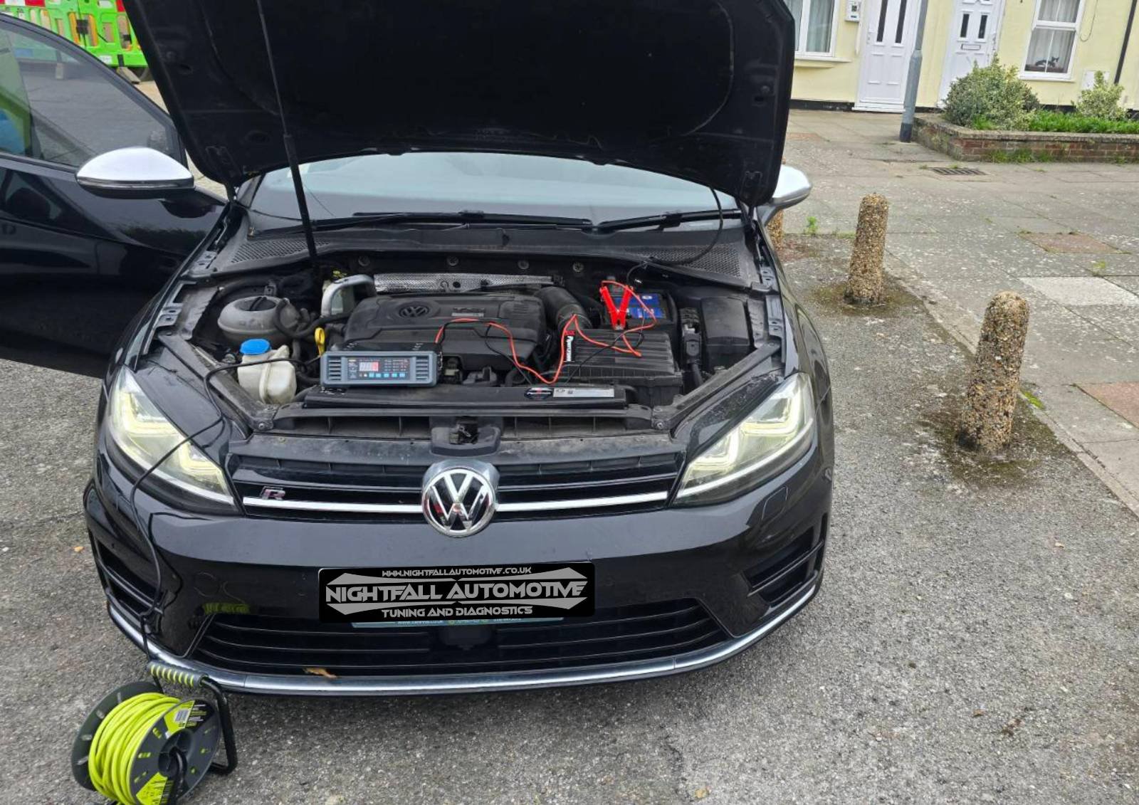 A black Volkswagen car with open hood showing engine and diagnostic equipment connected, parked on a street in front of pavement and small posts, with a yellow building in the background.
