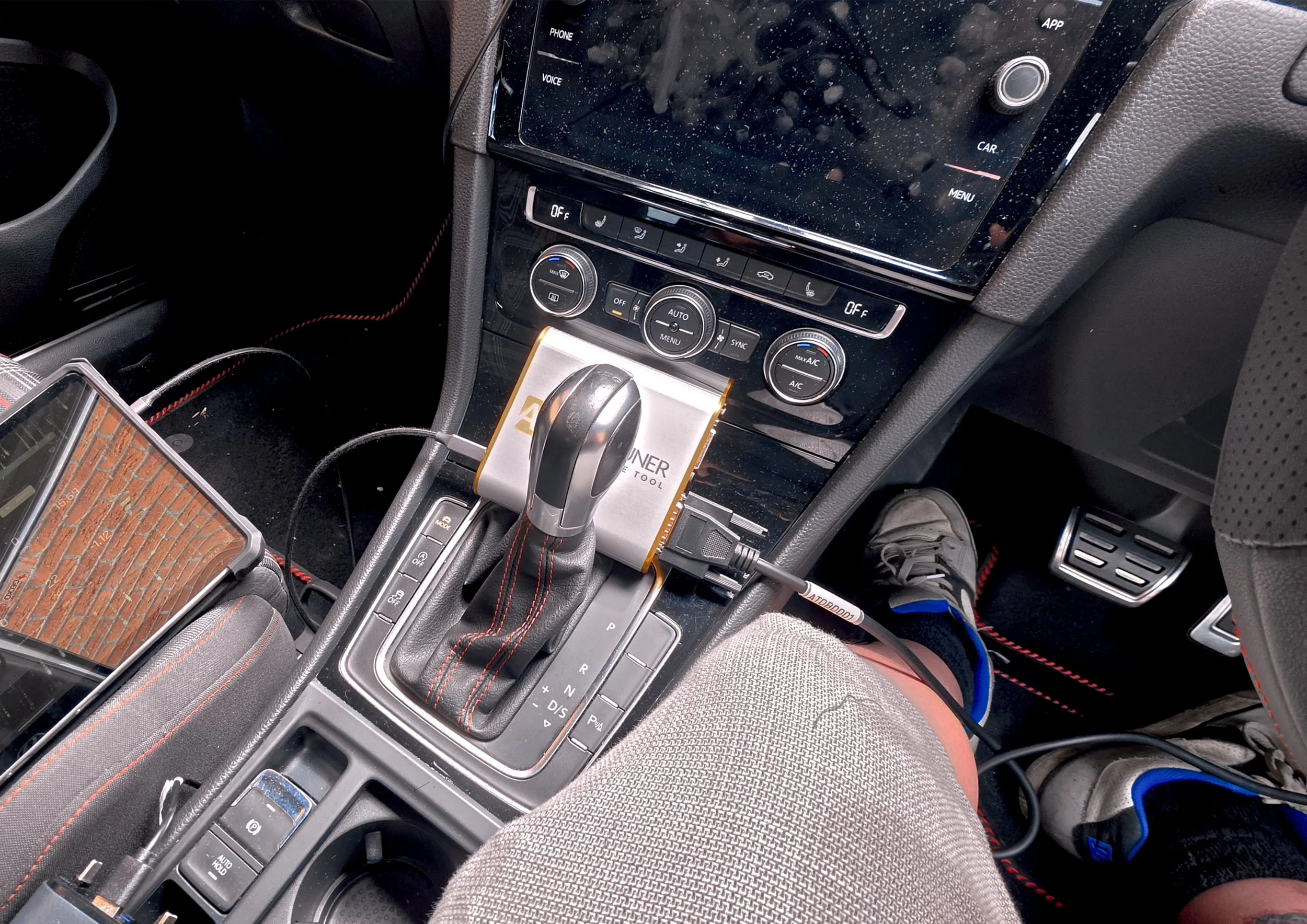 The interior of a car showing the center console with controls for climate and media, gear shift, and parts of the dashboard, with visible leg and foot of a passenger wearing gray shorts and sneakers. tuned by Nightfall Automotive using Autotuner